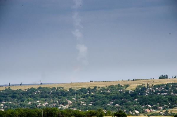 A vertical photograph taken from a residential area in Torez, Ukraine, showing a distinct, narrow white plume of smoke rising nearly vertically into a grey, overcast sky. The smoke trail originates from behind a line of trees and rooftops in the foreground, marking the launch trajectory of a Buk missile.